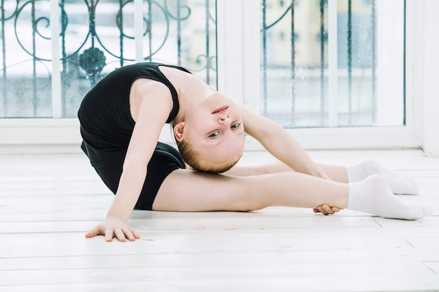 Piccolo ginnasta della ragazza del bambino che fa stretching in una stanza luminosa su uno sfondo della finestra felice e carino