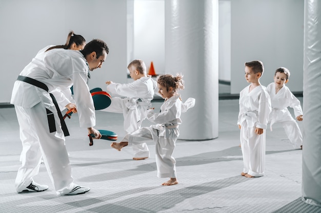 Piccolo gruppo di bambini in dobok che si esercitano con i loro allenatori di taekwondo mentre prendono a calci il bersaglio.