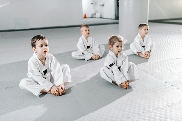 Piccolo gruppo di bambini sportivi caucasici che si siedono sul pavimento al taekwondo che prepara un ascolto al loro istruttore.