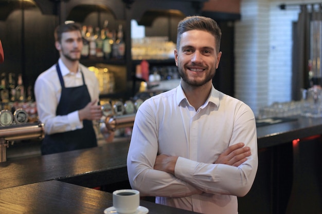 Piccolo imprenditore di successo in piedi con le braccia incrociate con un dipendente sullo sfondo che prepara il caffè.