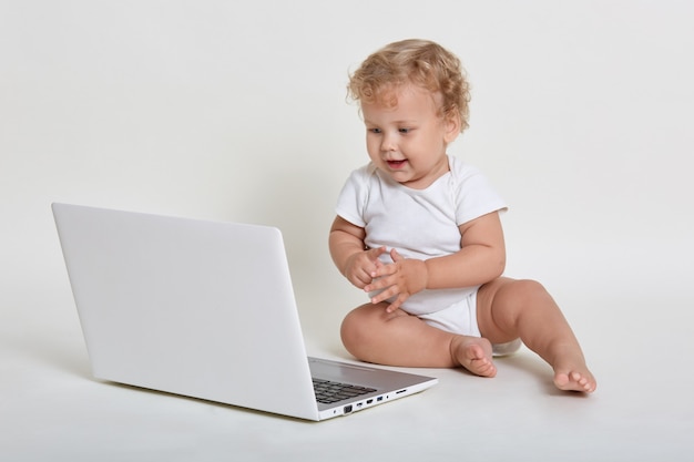 Piccolo maschio dai capelli ricci biondo che indossa il vestito del corpo, seduto sul pavimento con il lap top