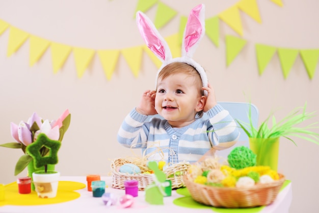 Piccolo neonato in orecchie di coniglio sorridente seduto al tavolo con decorazioni di Pasqua