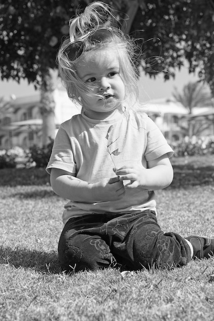 Piccolo neonato o bambino carino con viso adorabile e capelli biondi in camicia occhiali da sole e pantaloni seduti sull'erba verde giocando con foglie soleggiate all'aperto su sfondo naturale