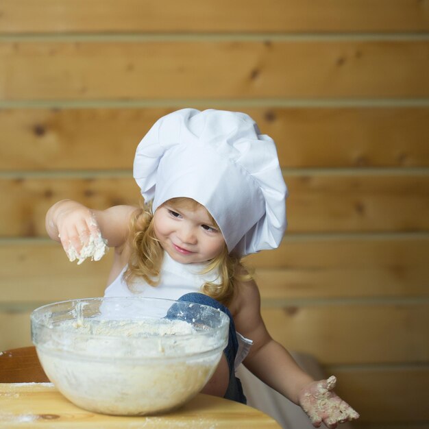 Piccolo neonato o bambino sveglio con la faccia felice in uniforme bianca del cuoco con il cappello del cuoco unico e il grembiule che impastano la pasta con la farina in ciotola di vetro in cucina su fondo di legno o di legno