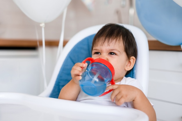 Piccolo neonato seduto nel seggiolone blu a casa sulla cucina bianca e acqua potabile dalla tazza sippy su sfondo con palloncini.