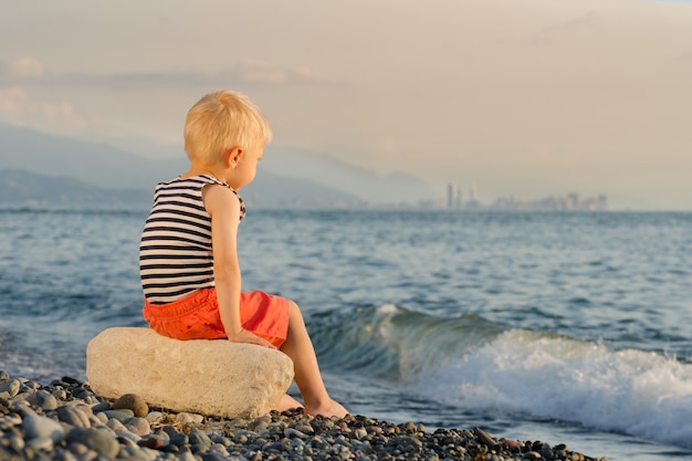 Piccolo ragazzo biondo in camicia a strisce che si siede sulla roccia sulla riva del mare. Bambino sulla spiaggia al tramonto