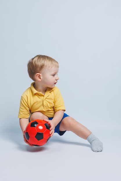 Piccolo ragazzo carino bambino ragazzo 34 anni tifoso di calcio in maglietta rossa tenendo in mano pallone da calcio isolato su sfondo giallo Bambini sport famiglia tempo libero stile di vita concetto Copia spazio pubblicitario