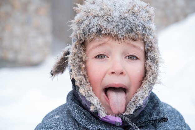 Piccolo ragazzo carino in cappello invernale innevato. Mostra la lingua.