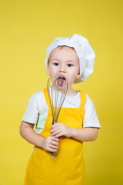 Piccolo ragazzo caucasico che gioca chef, ragazzo in grembiule e cappello da chef che tiene una frusta per montare la panna su sfondo giallo
