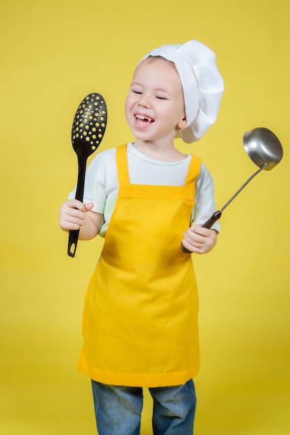 Piccolo ragazzo caucasico che gioca chef, ragazzo in grembiule e cappello da chef in posa emotivamente su sfondo giallo