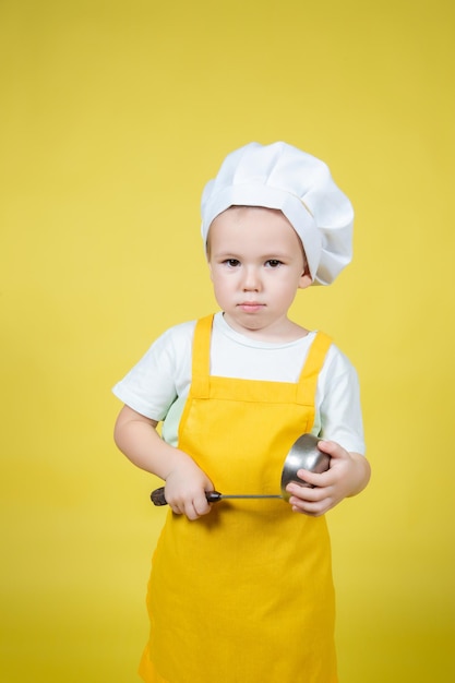 Piccolo ragazzo caucasico che gioca chef, ragazzo in grembiule e cappello da chef in posa su sfondo giallo