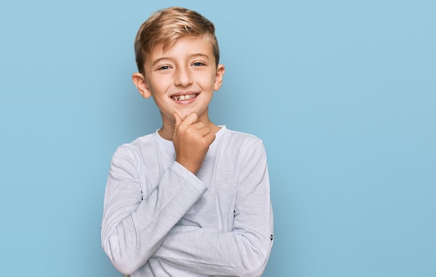 Piccolo ragazzo caucasico che indossa abiti casual che guarda fiducioso alla telecamera sorridendo con le braccia incrociate e la mano alzata sul mento. pensare positivo.