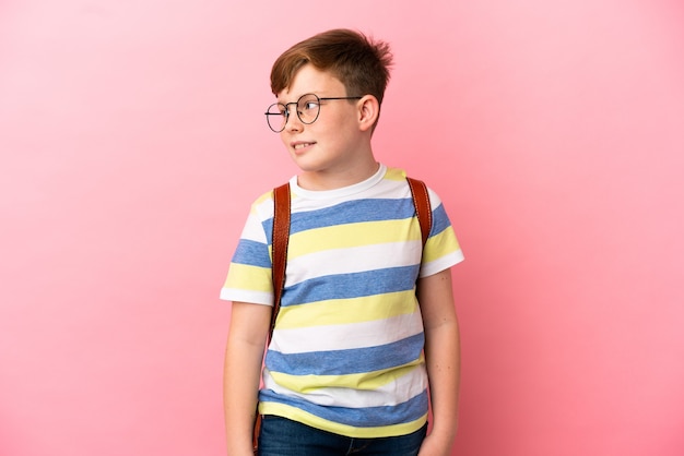 Piccolo ragazzo caucasico dai capelli rossi isolato su sfondo rosa guardando di lato e sorridente