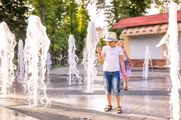 Piccolo ragazzo caucasico in cappello che gioca e che si diverte con acqua in fontana nel parco soleggiato di estate