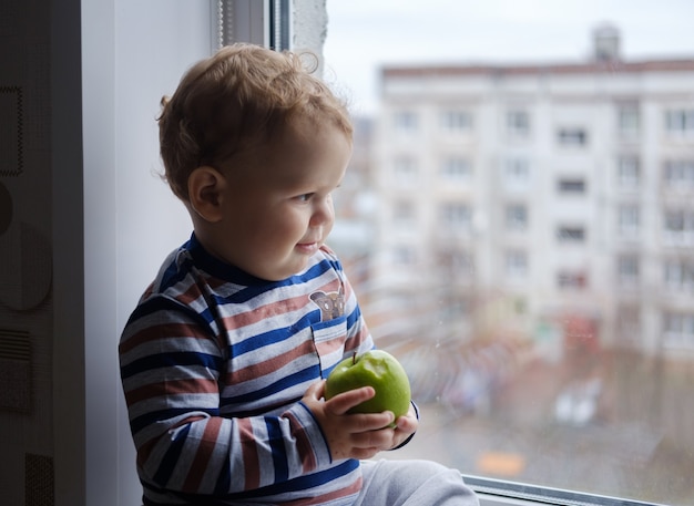 Piccolo ragazzo europeo mangia una mela verde seduta sul davanzale della finestra nella stanza