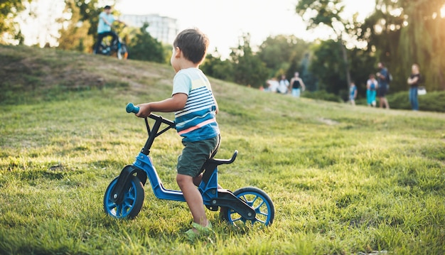 Piccolo ragazzo in sella a una bicicletta nel parco guardando altri bambini che camminano con i loro genitori
