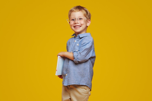Piccolo scolaretto intelligente che indossa occhiali da nerd e camicia blu che tiene blocchi di appunti e sorride alla telecamera