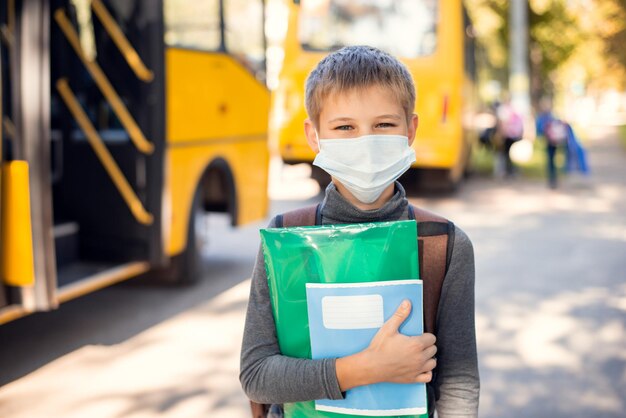 Piccolo scolaro con maschera medica che tiene in mano materiali didattici in piedi vicino a uno scuolabus