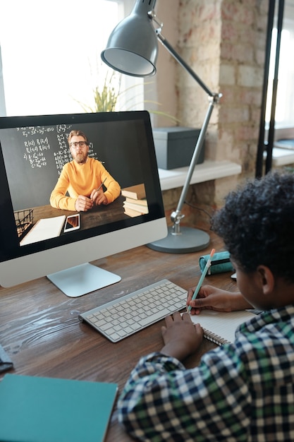 Piccolo scolaro contemporaneo seduto al tavolo davanti al monitor del computer durante la lezione online, ascoltando il suo insegnante sullo schermo e prendendo appunti