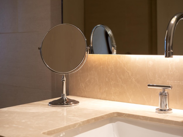 Piccolo specchio per il trucco rotondo pieghevole in piedi vicino al lavabo con piano in marmo e rubinetto in acciaio vicino alla grande finestra in vetro con illuminazione nascosta Design moderno di interni bianchi e puliti del bagno