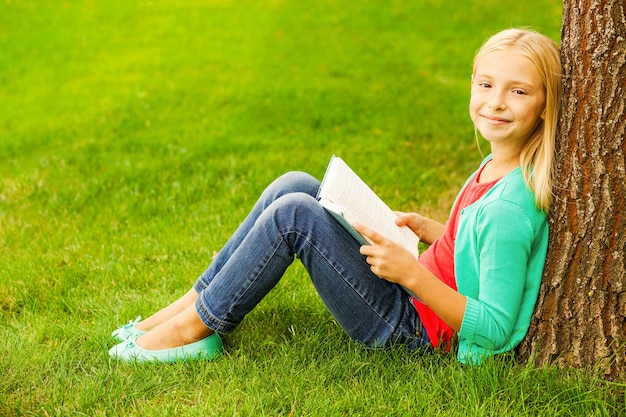 Piccolo topo di biblioteca. Ragazza carina con i capelli biondi che tiene in mano un libro e sorride mentre è seduta sull'erba verde e si appoggia all'albero