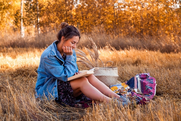 Picnic all'aria aperta: una giovane donna in giacca di jeans e vestito legge libri sul plaid con un cestino da picnic, mele, vino.