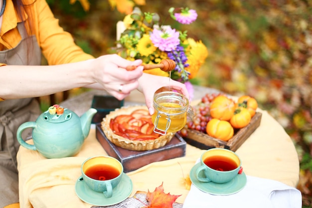 Picnic autunnale Donna in abito giallo e grembiule di lino beve il tè dalla tazza al tavolo di legno in giardino Bella tovaglia bollitore miele con cucchiaio torta di mele raccolta cachi uva foglia d'acero