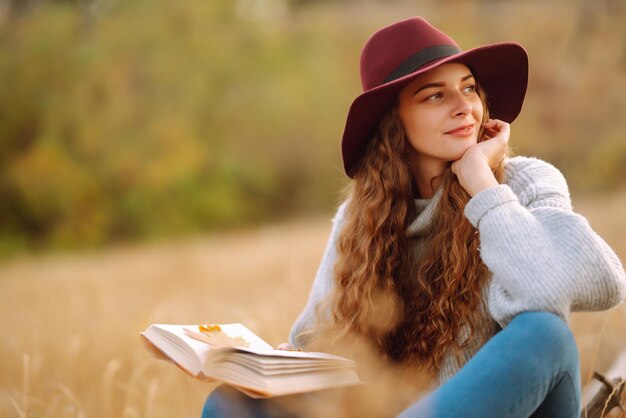 Picnic autunnale in natura Giovane donna seduta sulla coperta leggendo un libro e sorridendo all'aperto