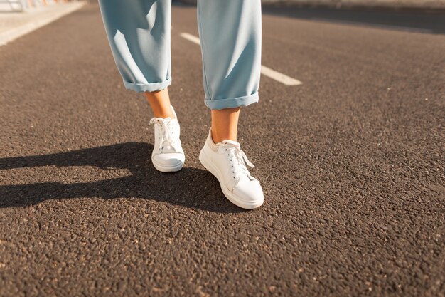 Piedini femminili alla moda del primo piano in pantaloni blu alla moda in scarpe da ginnastica alla moda bianche stanno sull'asfalto in una luminosa giornata di sole. Collezione estiva moderna alla moda di scarpe da donna. Stile casual.