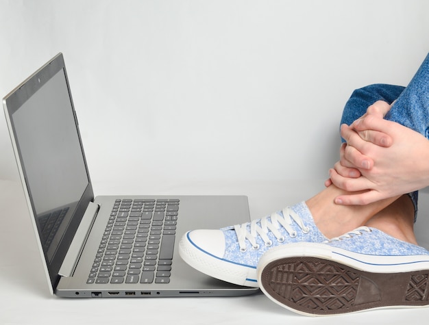 Piedini femminili in jeans, scarpe da ginnastica e un computer portatile su un bianco.