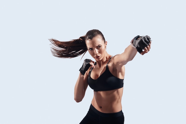 Pieno di forza. Attraente giovane donna che guarda la macchina fotografica e fa la boxe in piedi su sfondo grigio