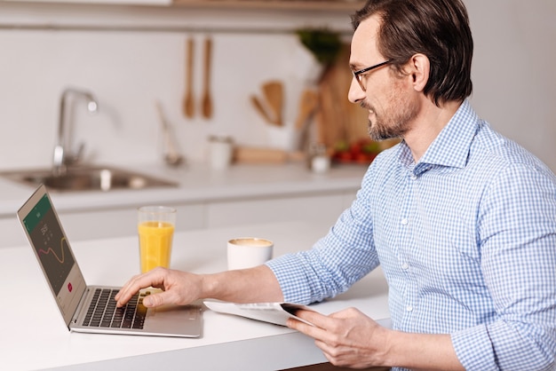 Pieno di ispirazione. Sorridente soddisfatto sincero imprenditore autonomo seduto a casa e durante l'utilizzo di laptop e la lettura di nuovi