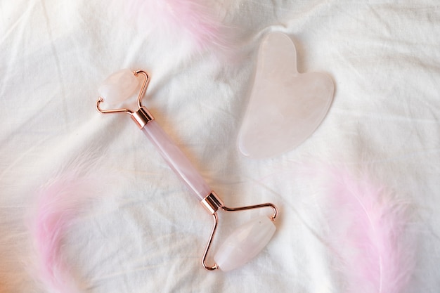 Pietra di quarzo naturale rosa gua sha e strumento di massaggio a rullo per massaggio al viso decorato con piume rosa su fondo di cotone bianco. Concetto di bellezza con accessorio alla moda