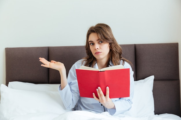 Pigiama d'uso della giovane donna confusa e libro della tenuta a letto