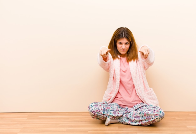 Pigiama da portare della giovane donna che si siede a casa che indica in avanti alla macchina fotografica con entrambe le dita e l'espressione arrabbiata, dicendovi di fare il vostro dovere