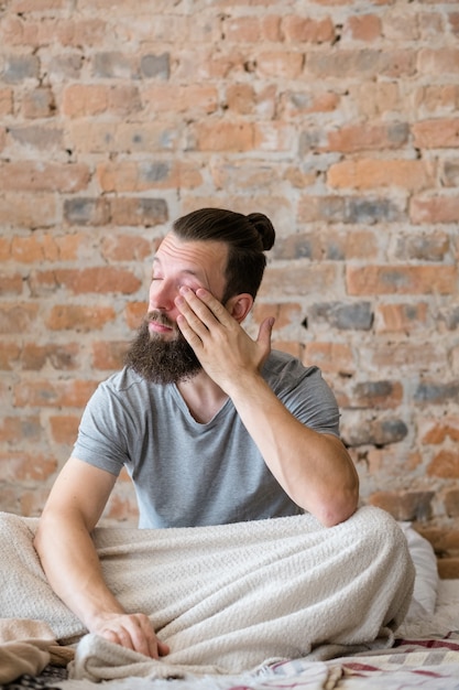 Pigrizia mattutina e stanchezza. Cattiva notte di sonno. Uomo sonnolento seduto a letto e strofinando gli occhi. Copia spazio.