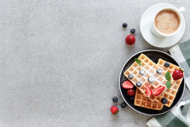 Pila di cialde in polvere di zucchero con miele su un piatto sull'asciugamano e cemento grigio o tavolo in ardesia con mirtillo, fragola tritata e foglie di menta e tazza bianca e piatto di caffè