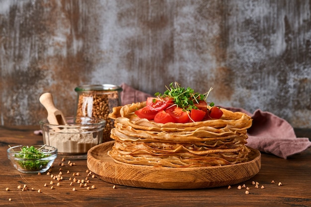 Pila di crepes di farina di grano saraceno senza glutine frittelle con pomodorini e microgreens di rucola su piatto di legno, cottura sana fatta in casa per la colazione