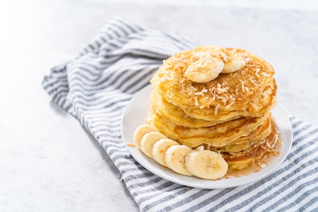 Pila di frittelle di banana al cocco appena sfornate guarnite con banane a fette, cocco tostato e sciroppo d'acero.