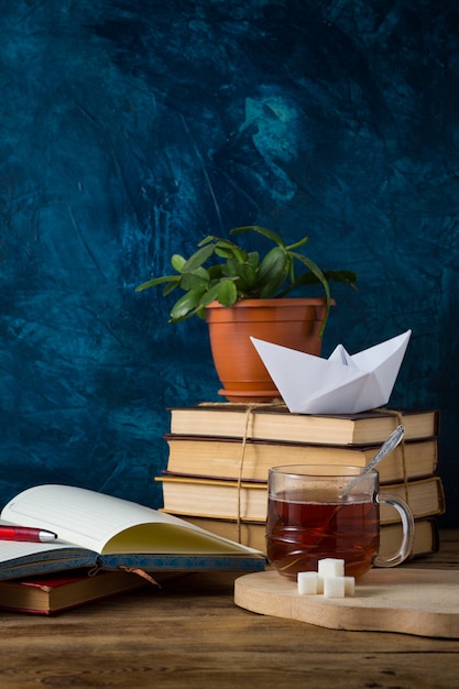 Pila di libri, una tazza di tè caldo, una barchetta di carta, un diario aperto con una penna su un muro blu scuro