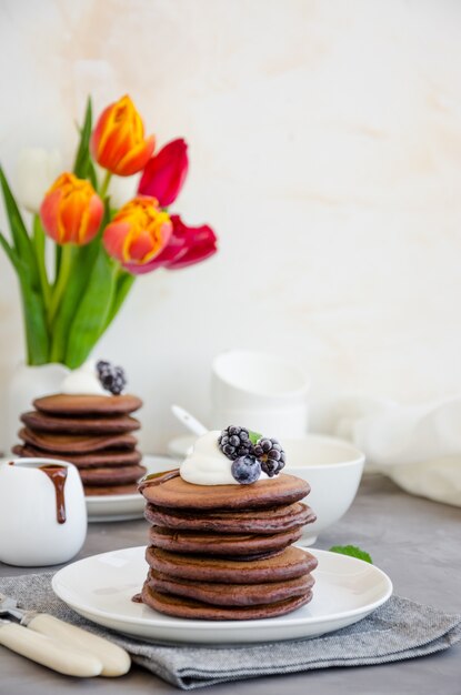 Pila di pancake al cioccolato su un piatto bianco con salsa di cioccolato, panna montata, mirtilli e more in cima a uno sfondo concreto.