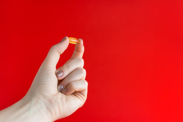 Pillola gialla tra due dita. La mano di una ragazza tiene una pillola trasparente su sfondo rosso. Capsule d'oro di olio di pesce, olio di pesce omega-3 nella palma femminile