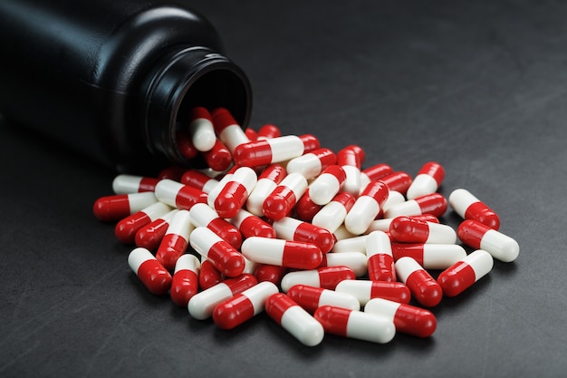 Pillole rosse e bianche con capsule versate da un barattolo nero su sfondo nero.