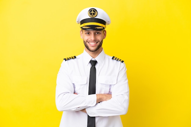 Pilota caucasico dell'aeroplano isolato su sfondo giallo mantenendo le braccia incrociate in posizione frontale