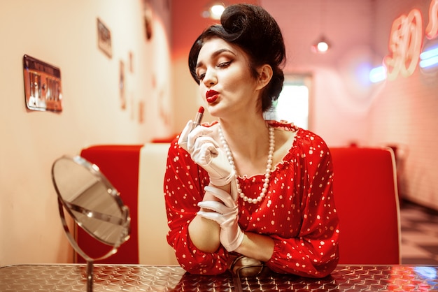 pin up donna con rossetto luminoso in mano seduto contro lo specchio, vestito con pois, stile vintage. Moda americana retrò cafe 50