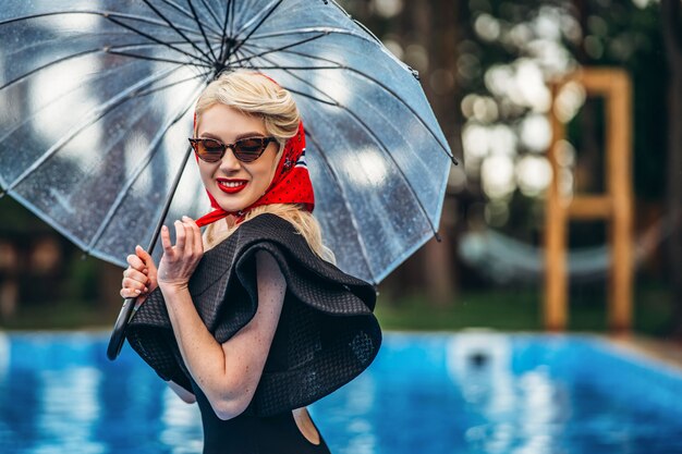 Pin up in stile bionda in occhiali da sole tenendo l'ombrello e divertirsi in piscina