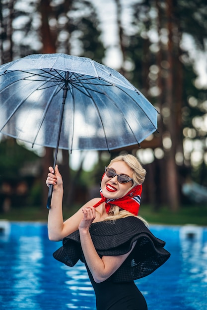 Pin up in stile bionda in occhiali da sole tenendo l'ombrello e divertirsi in piscina