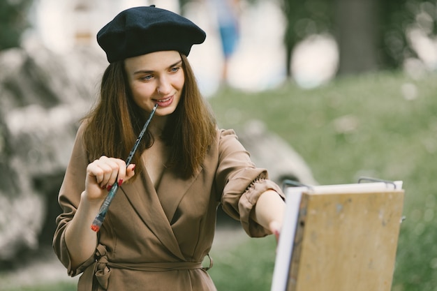 Pittore femminile che dipinge nel parco. Una donna che indossa un abito color cachi e un cappello nero