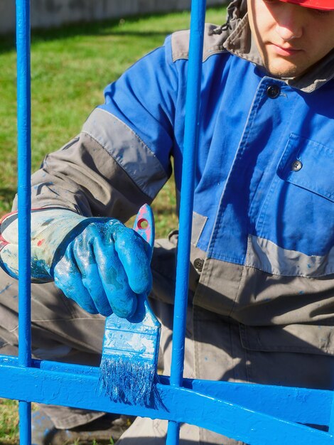 Pittore professionista in abbigliamento e casco dipinge recinzione metallica o recinzione fatta di rinforzo con pennello il giorno d'estate Flusso di lavoro autentico Foto di lavoro reale