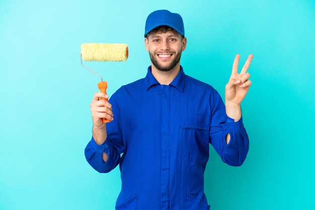 Pittore uomo caucasico isolato su sfondo blu sorridente e mostrando il segno di vittoria